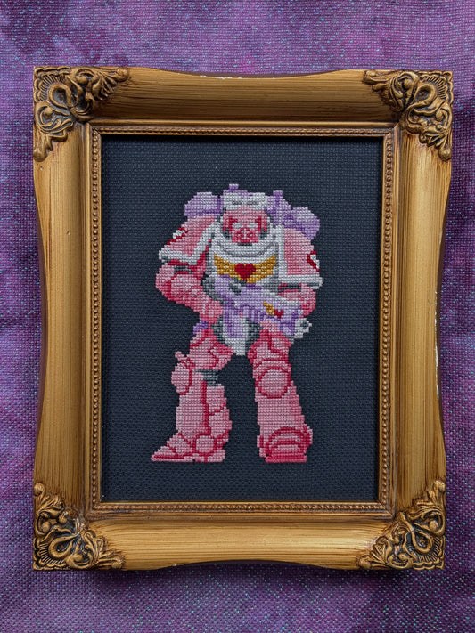 A framed cross-stitch artwork featuring a pink and purple pastel space marine on a black fabric background, enclosed in an ornate gold frame against a purple textured backdrop.