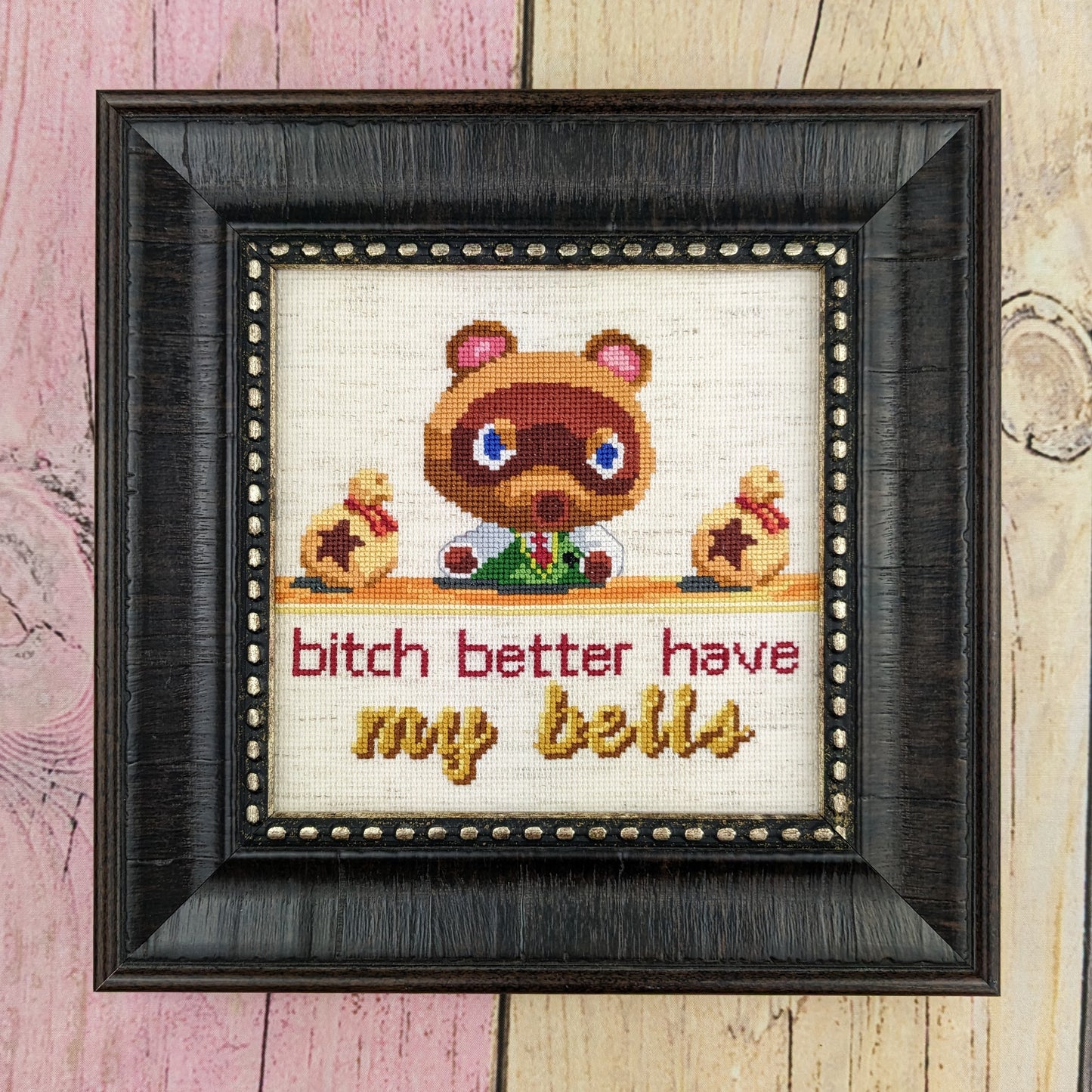 A cross stitch depicting Tom Nook from Animal Crossing New Horizons sitting at his desk with an angry look on his face. He is flanked by two bags of bells laying on his desk. Text under the desk reads "bitch better have my bells", with the words "my bells" being cursive and golden. The stitch is done on oatmeal aida and displayed in a black square frame on pastel stained fenced wood.