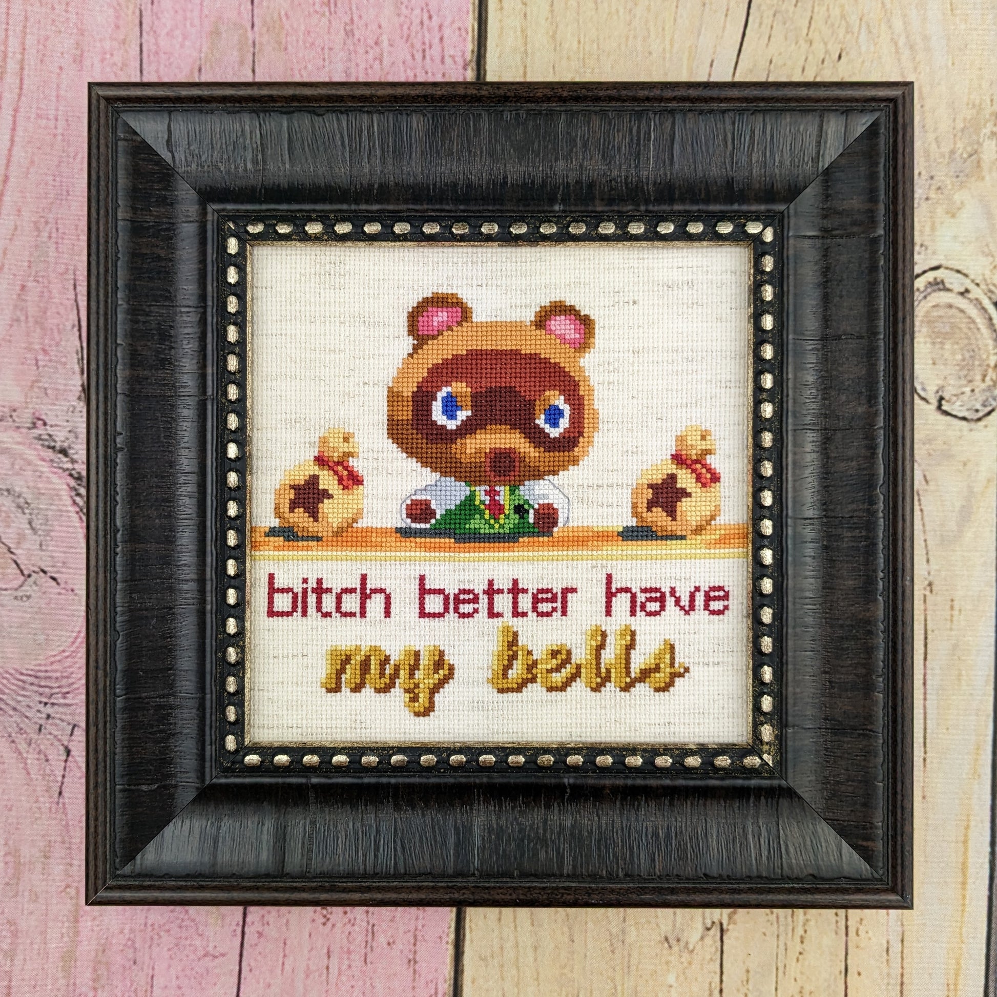A cross stitch depicting Tom Nook from Animal Crossing New Horizons sitting at his desk with an angry look on his face. He is flanked by two bags of bells laying on his desk. Text under the desk reads "bitch better have my bells", with the words "my bells" being cursive and golden. The stitch is done on oatmeal aida and displayed in a black square frame on pastel stained fenced wood.