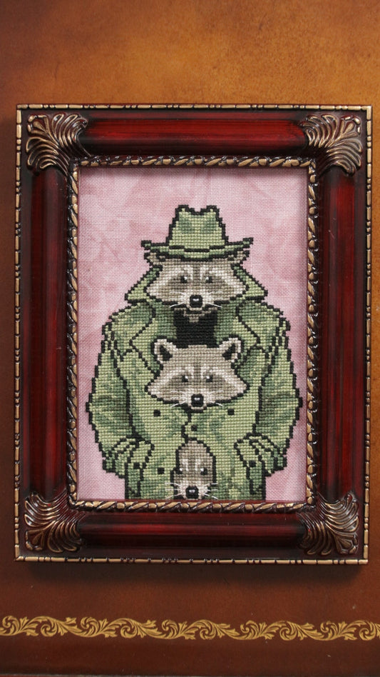 Framed cross-stitched picture of three raccoons in a green trench coat, wearing a matching fedora on a pink background
