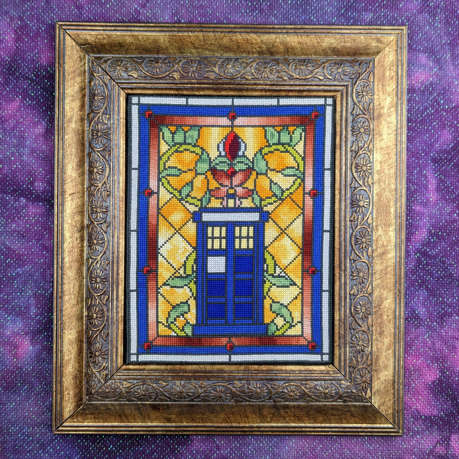 A picture of a cross stitch in an elaborate gold frame. The cross stitch imitates the look of a stained glass window with a TARDIS in the center of the frame, surrounded by vines.