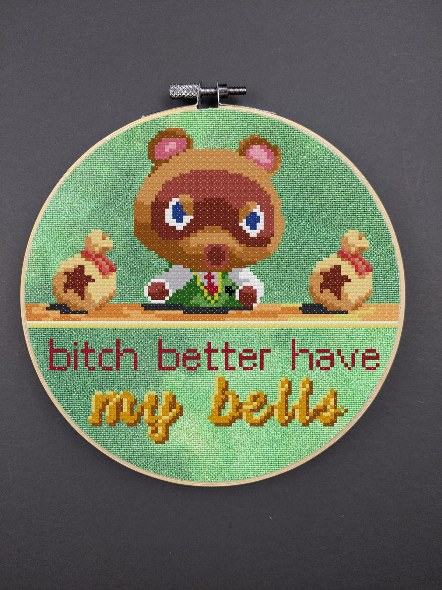 A digital rendering of a cross stitch depicting Tom Nook from Animal Crossing New Horizons sitting at his desk with an angry look on his face. He is flanked by two bags of bells laying on his desk. Text under the desk reads "bitch better have my bells", with the words "my bells" being cursive and golden.  The stitch is depicted on a green hand-dyed opalescent aida and displayed in a hoop on a gray background.